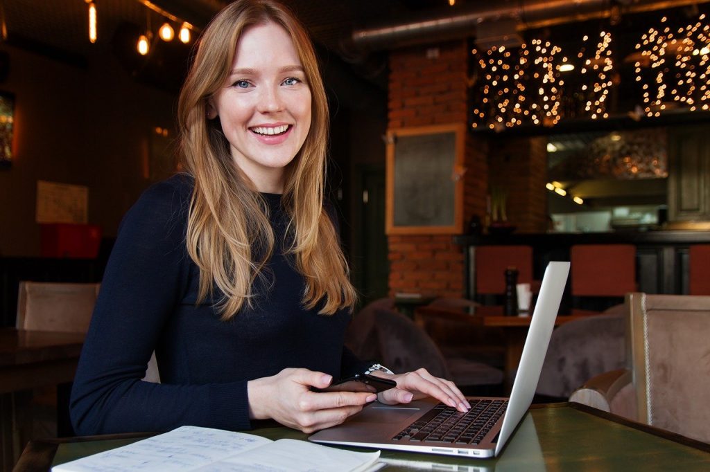 Nuori punatukkainen nainen hymyilee ja käyttää puhelinta käsi tietokoneen näppämistöllä.